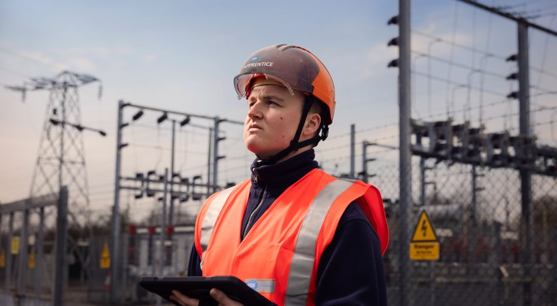 Apprentice technician holding a tablet, standing near electrical station