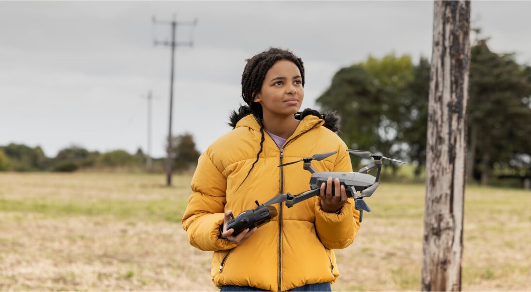 Teen girl looking puzzled with a drone in her hands, standing in a field with electric poles around here.