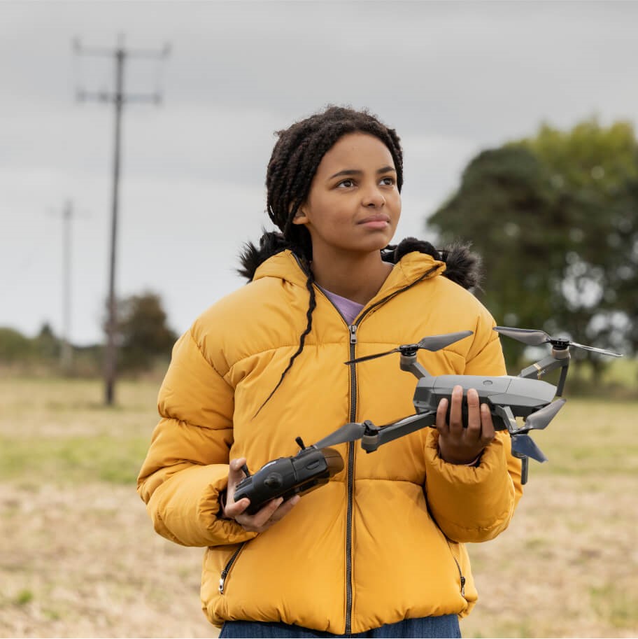 Teen girl looking puzzled with a drone in her hands, standing in a field with electric poles around here.