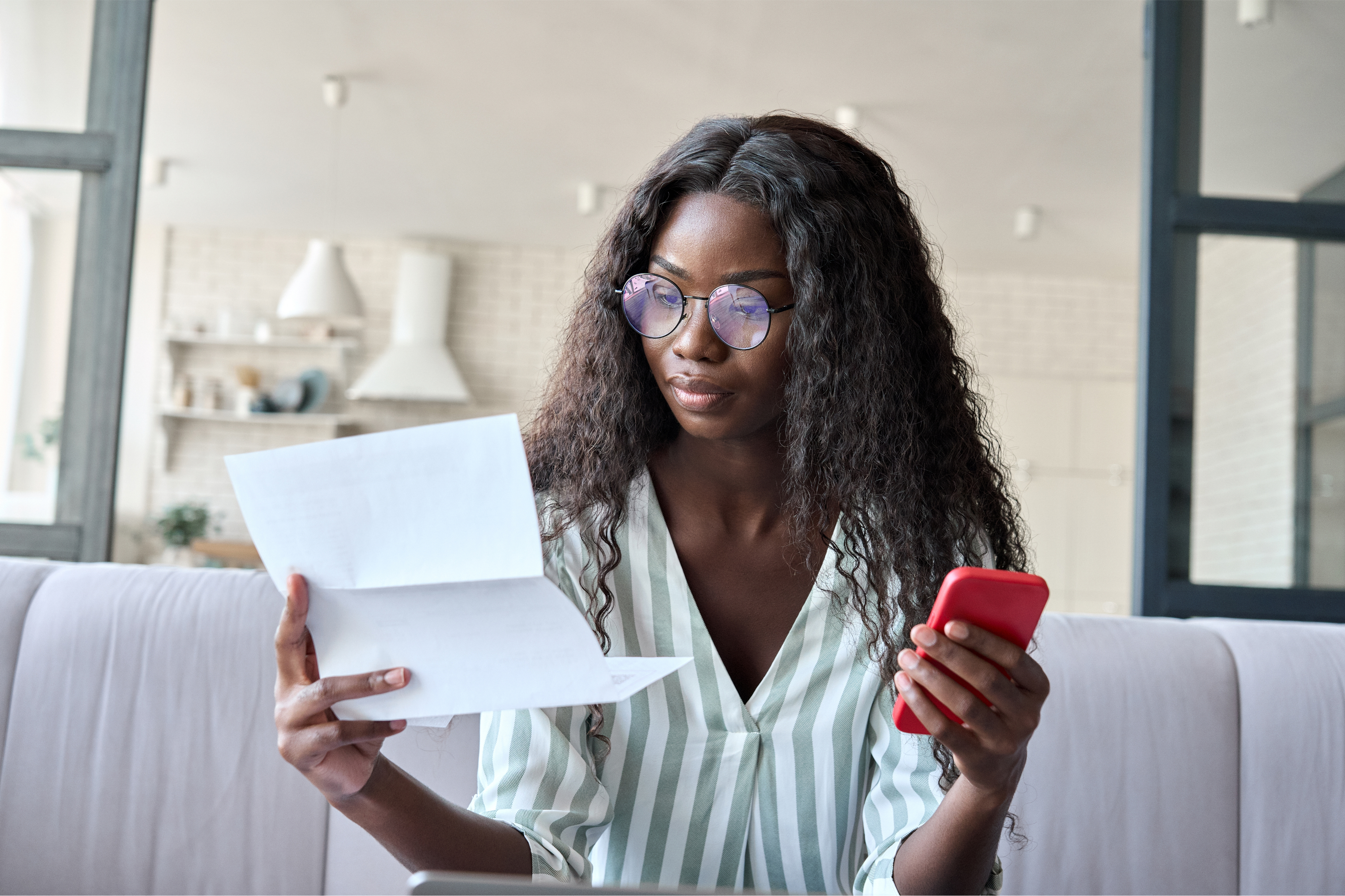 A woman with curly hair and glasses reading paper, holding her smartphone.